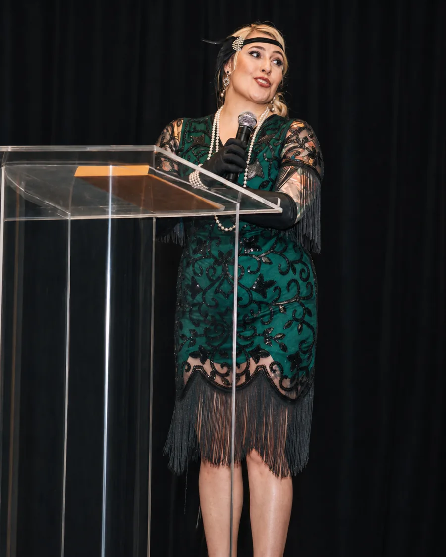 Janelle Nightingale hosting at an acrylic podium in evening dress, holding a microphone.