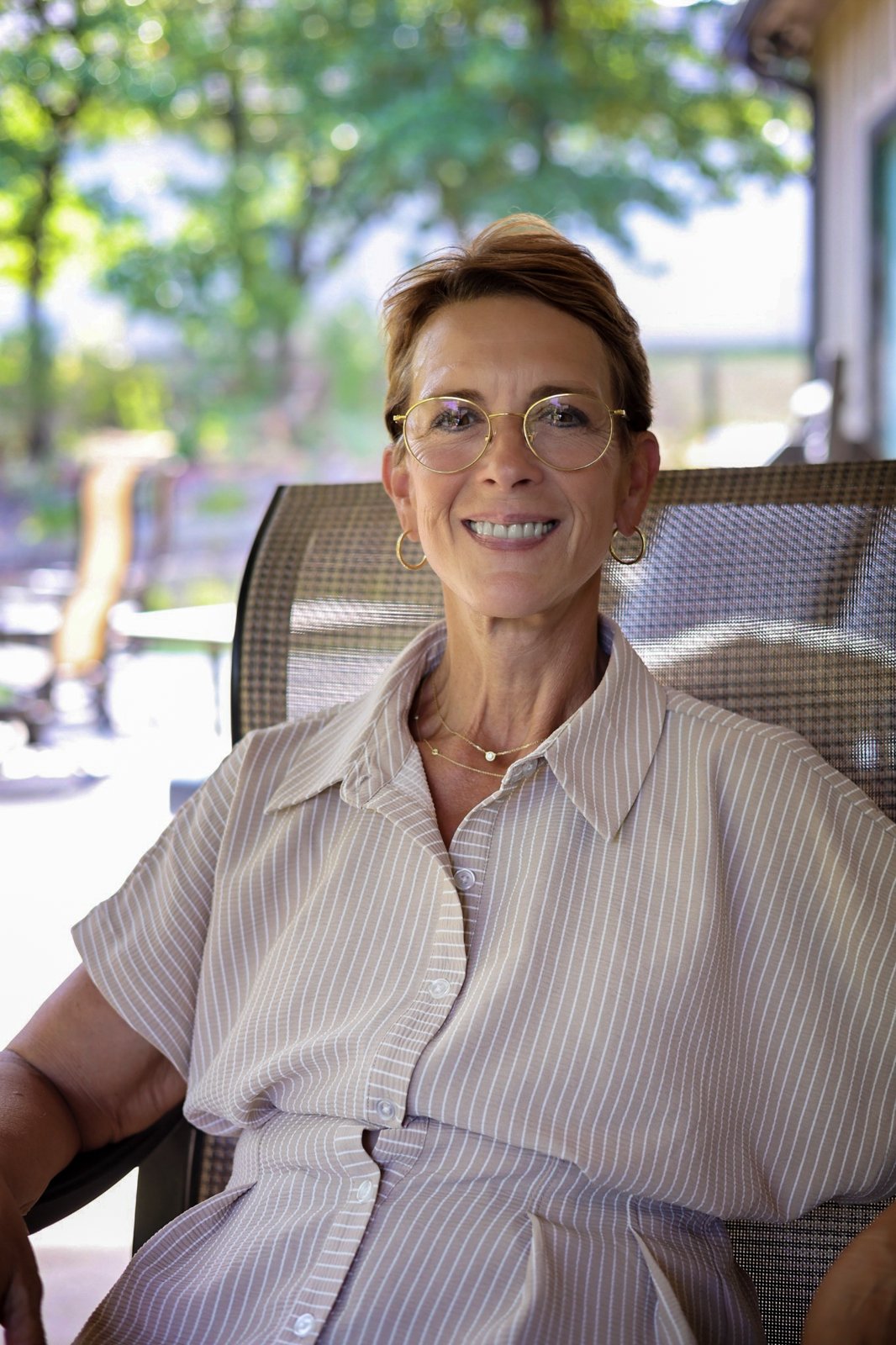 A smiling woman wearing round glasses and a striped shirt sits in a mesh chair on a shaded porch.