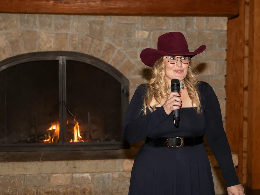 Janelle Nightingale speaking with a handheld microphone beside a lit stone fireplace.
