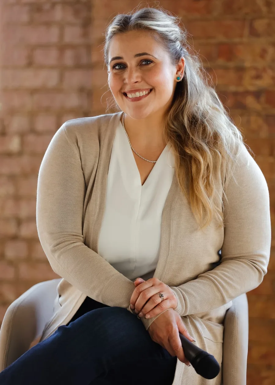 Janelle Nightingale seated and smiling in a linen cardigan against an exposed brick wall, holding a microphone.