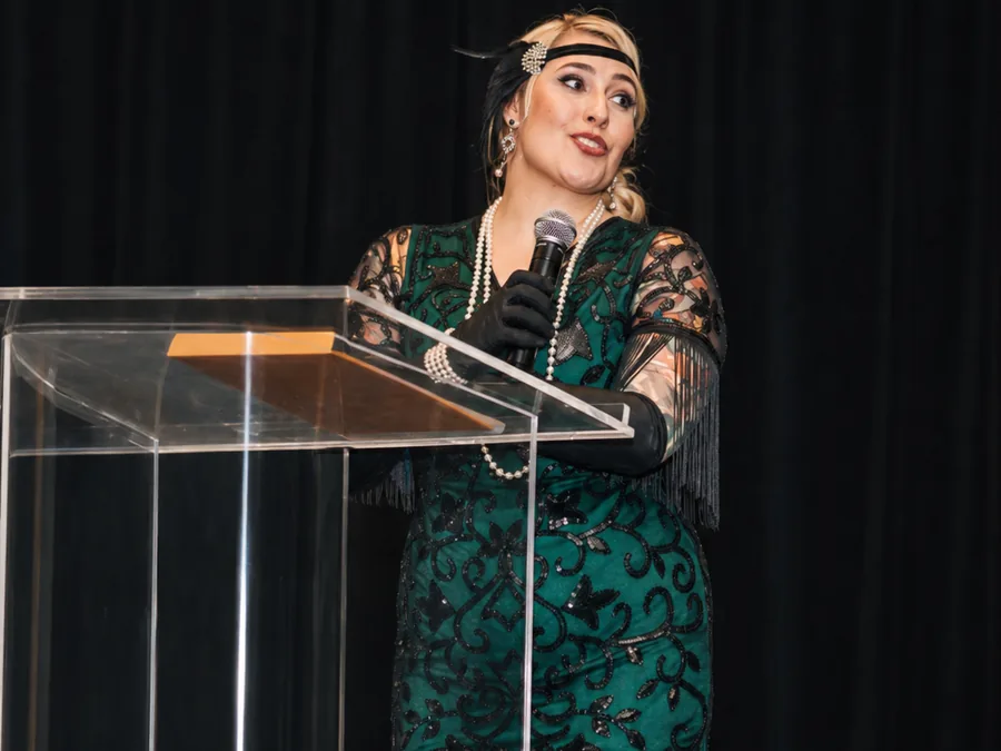 Janelle Nightingale hosting at an acrylic podium in evening dress, holding a microphone.