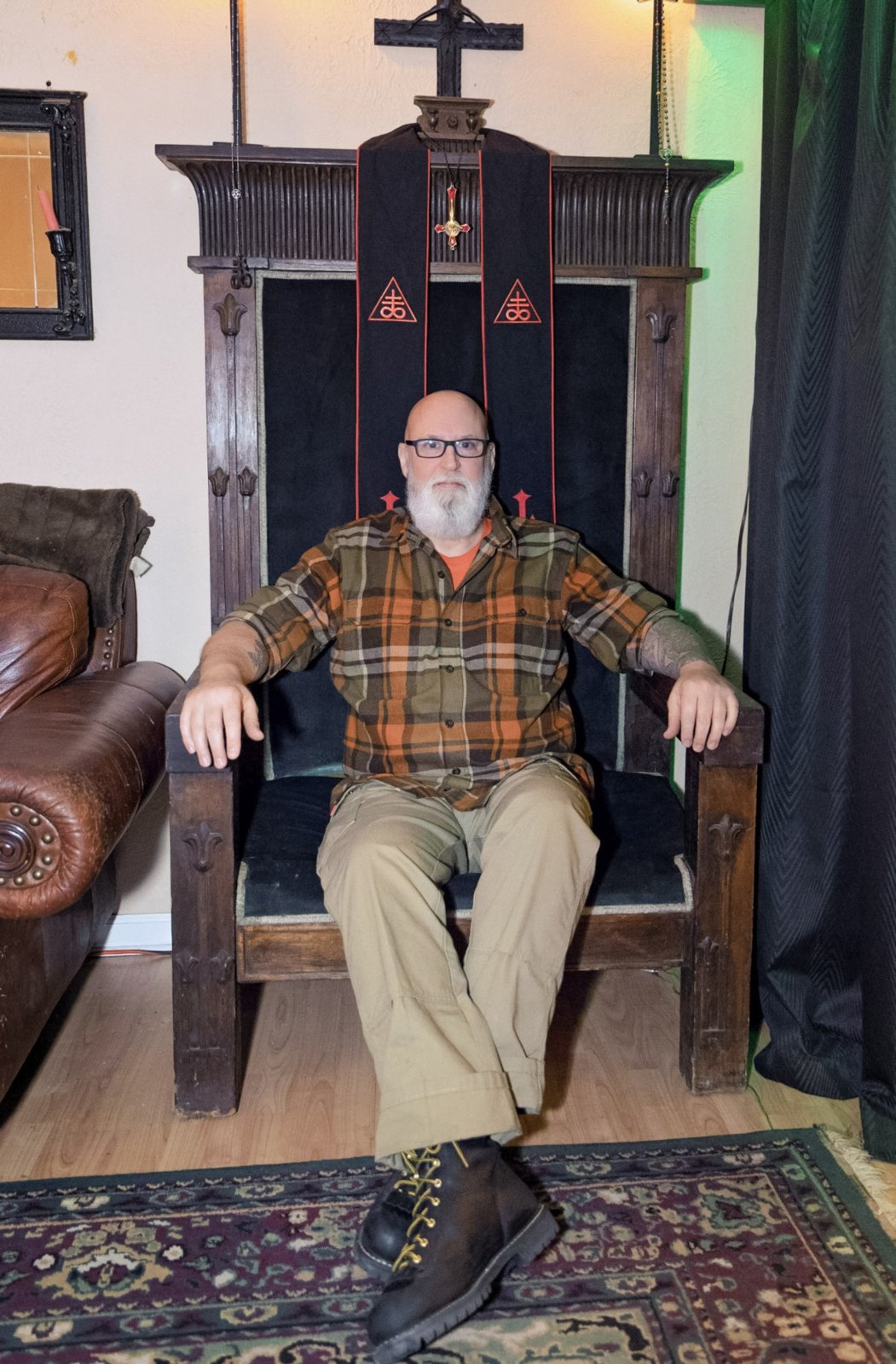 A bearded man wearing glasses and a plaid shirt sits in a tall wooden chair with his legs crossed.