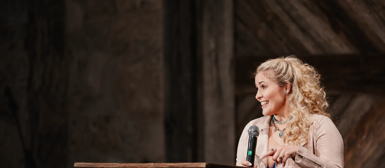 Janelle Nightingale speaking at a wooden podium with a handheld microphone, mid-gesture, in a barn venue.