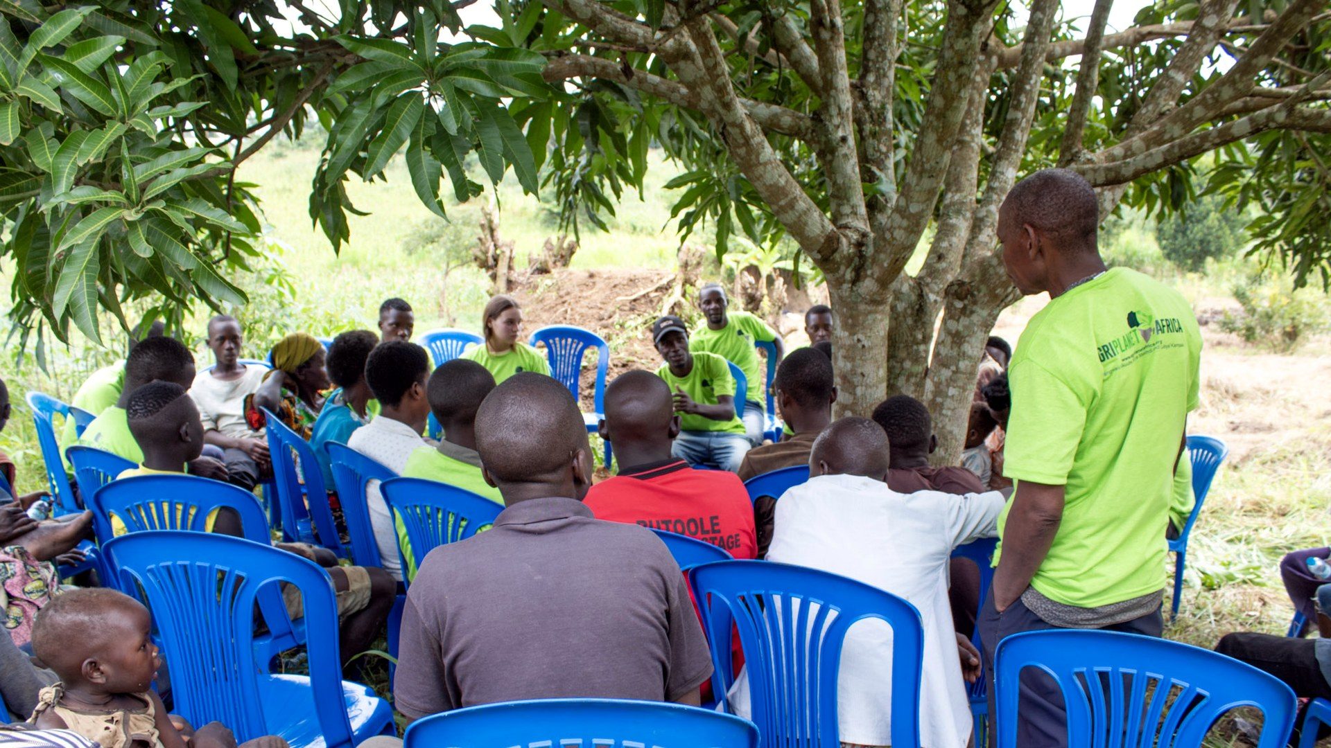 Community members and Agri Planet Africa staff sit in a wide circle of blue plastic chairs under a mango tree in Uganda, listening as one man speaks.