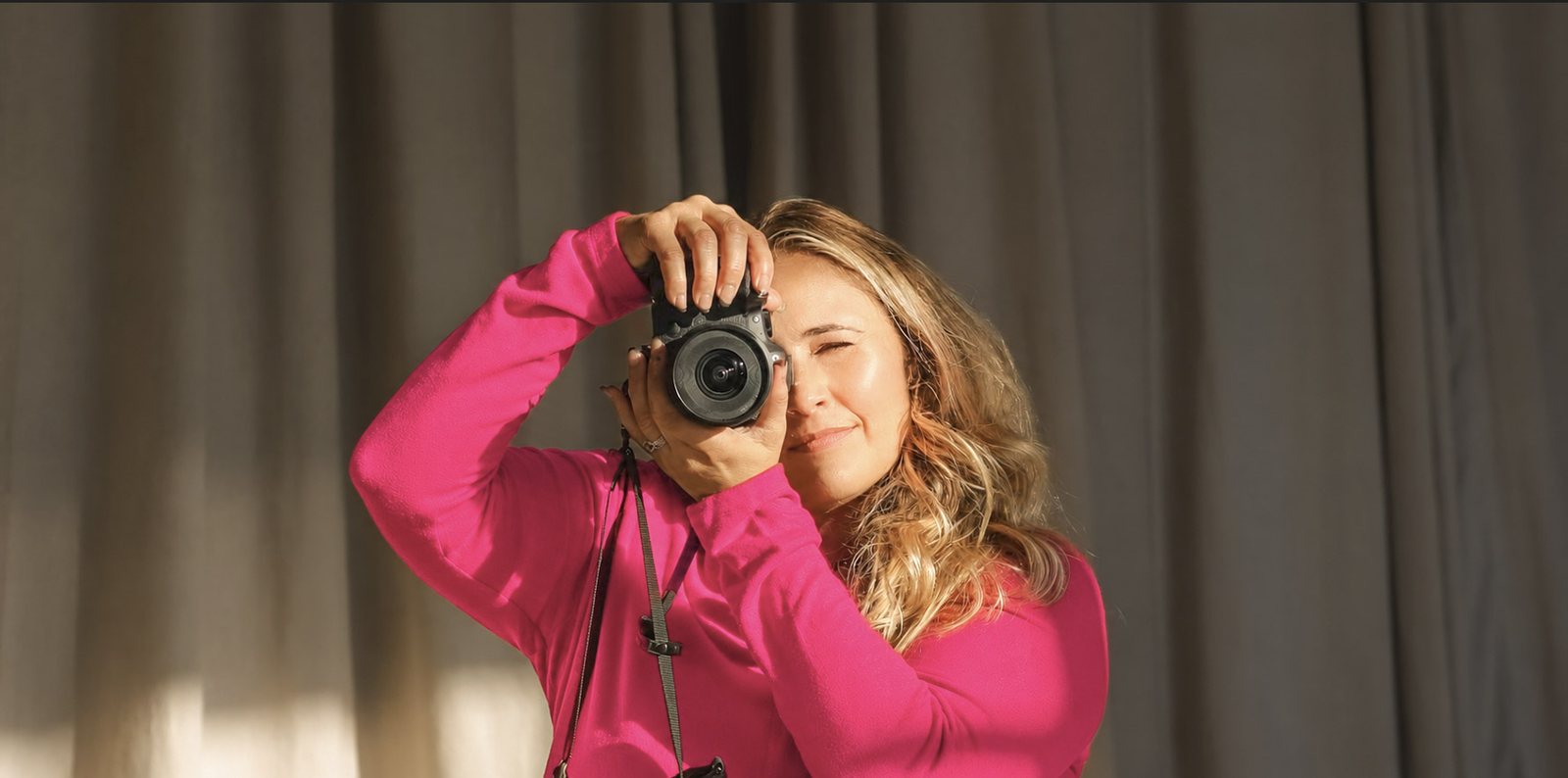 A woman in a pink sweater raises a camera to her eye against a curtain backdrop.