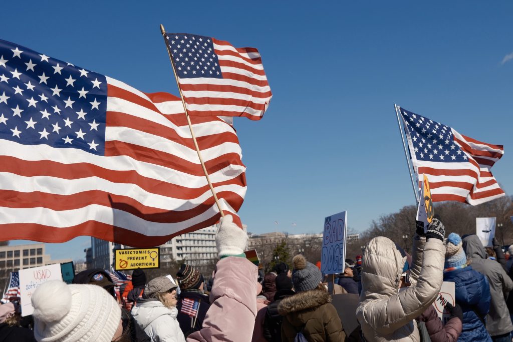 A group of protesters raises American flags in the air at the 50501 protest in Washington, D.C., on February 17, 2025. Demonstrators in warm winter clothing carry signs, including one that reads "Vasectomies prevent abortions" and another that says "No! Bad Doge". The energy of the protest is captured in this moment. Photo by Chance Clark.