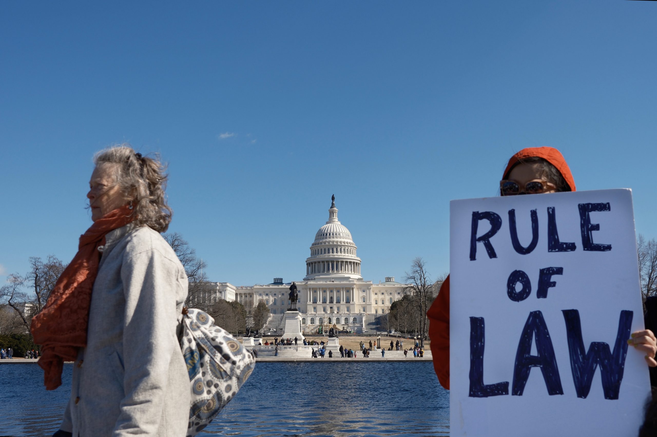 A protester holds a sign that reads "RULE OF LAW" in front of the U.S. Capitol during the 50501 protest in Washington, D.C., on February 17, 2025. Another protester walks past, wrapped in a scarf, highlighting the diverse group of demonstrators at the event. Photo by Chance Clark.