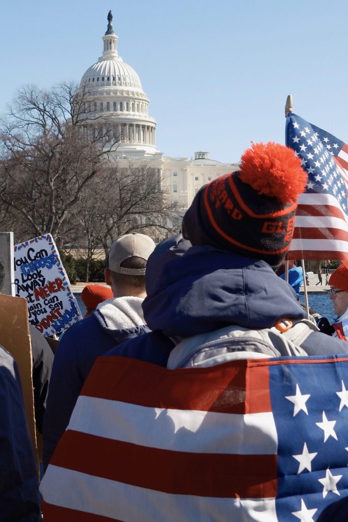 A protester wearing a navy hoodie and a backpack stands among a crowd at the 50501 protest in Washington, D.C., on February 17, 2025. The U.S. Capitol is visible in the background as demonstrators carry signs, including one that reads, "You can CHOOSE to look away but NEVER again can you say you DIDN'T KNOW." Photo by Chance Clark.