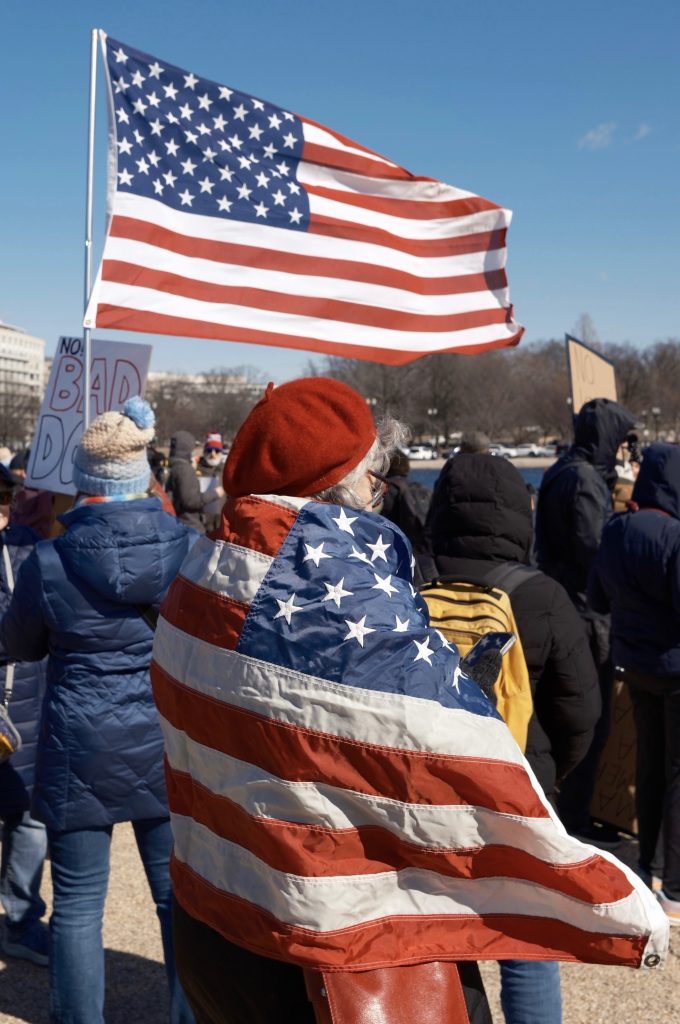 A woman in a red beret, wrapped in an American flag, stands among protesters at the 50501 protest in Washington, D.C., on February 17, 2025. A large American flag waves above her as demonstrators carry various signs. Photo by Chance Clark.